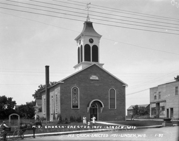 View across street of the exterior of the Methodist Evangelical Church, constructed in 1851. The building features a clock tower steeple, arched stained-glass windows, and a chimney. Two women and a child stand on the lawn in front of the church. A man in a buggy waits at the curb.