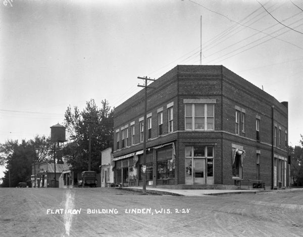 Wedge-shaped building on the corner of a commercial street. A water tower is located down the street. Caption reads: "Flatiron Building, Linden, Wis."