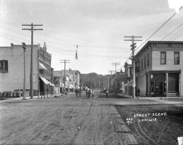 View down Main Street. The shops along the street include: Everson and Hinrich's grocery store, Perry Music House, and Bon Ton Restaurant. A man poses with his two horses in the center of the street.
