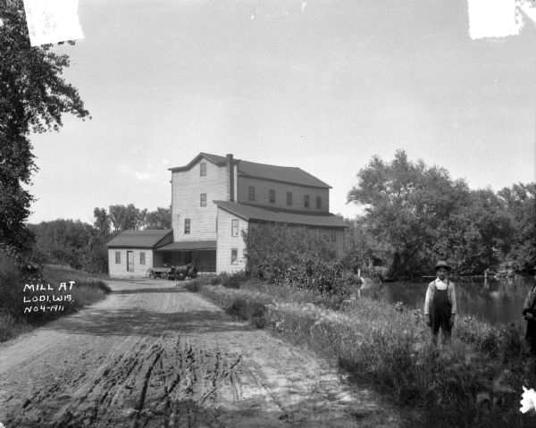 View down an unpaved road towards a four-story mill. Two boys pose on the side of the road.