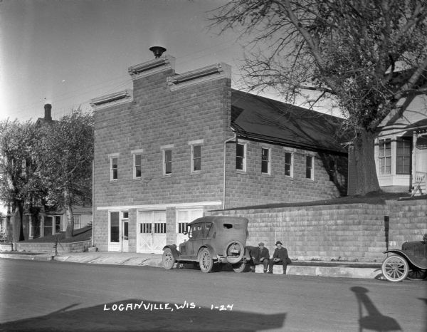 View across street of two men sitting on the street curb near a parked car. There is a garage on the left.