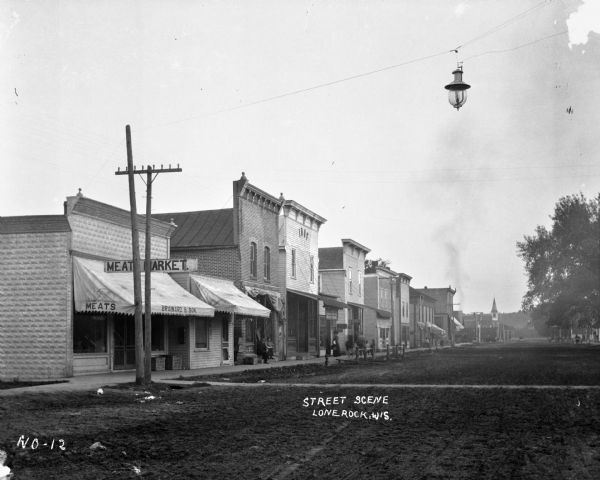 View across street of row of shops along one side of a main street. Brainard and Son Meat Market is located on the far left.