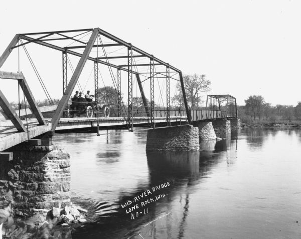View from shoreline of three people in a car on a bridge over the Wisconsin River.
