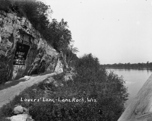 A pedestrian path along the Wisconsin River. An advertisement for J.M. Brophy general store is painted on the rock formations along the path.