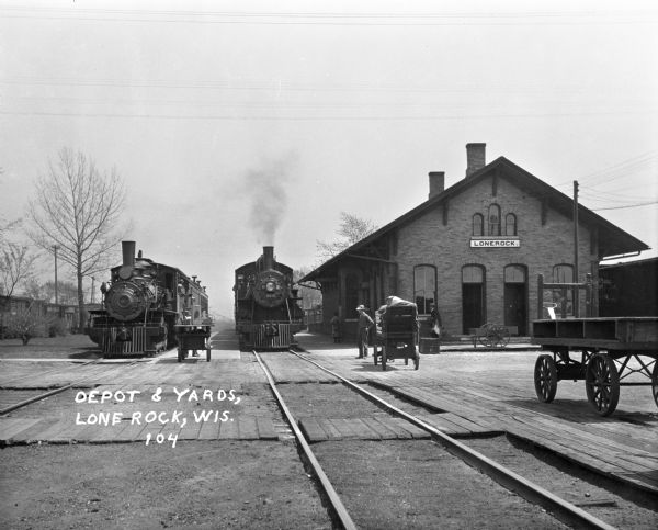Two trains at the Lone Rock depot. The platform holds waiting passengers and carts.