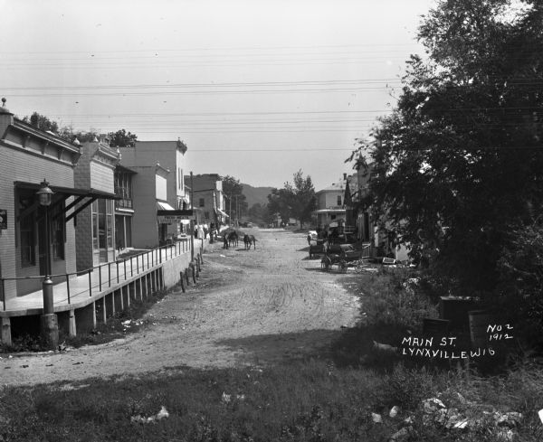 View down unpaved Main Street. People pose on the raised sidewalk near a shop selling "Fresh Meats" and "Groceries" on the left. Another group of people pose outside of a livery on the right. There are horses and carriages in street.