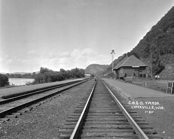 View down railroad tracks of the Lynxville train depot on the Chicago Burlington and Quincy Railroad line. There are bluffs on the right, and a man is walking down the railroad tracks. Two other men are walking near the depot.