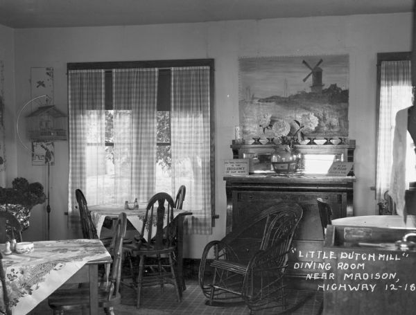 The dining room at the Little Dutch Mill Restaurant. The room has tables and chairs, a rocking chair, a bird cage, and signs that read, "Our only advertising is the excellency of our work" and "We aim to please - give us a trail."