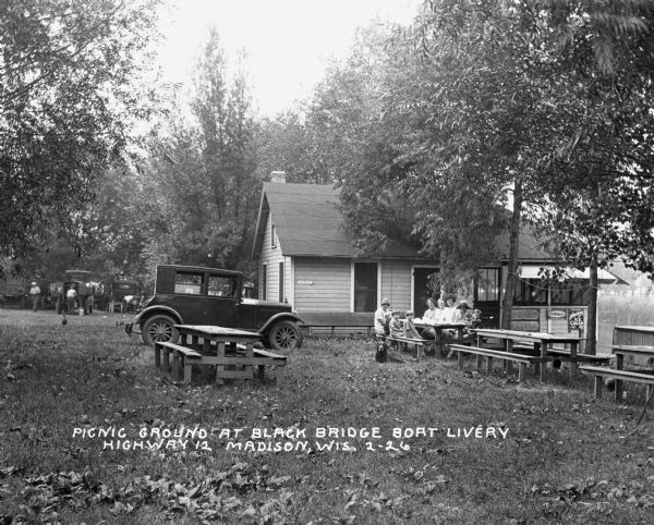 A group of women and children sit at a picnic table at the Black Bridge Boat Livery off Highway 12. Behind them is a building with a screened porch facing the water. A car is parked nearby, and a dog sits in the grass near the table. In the far background men stand near automobiles parked near trees.