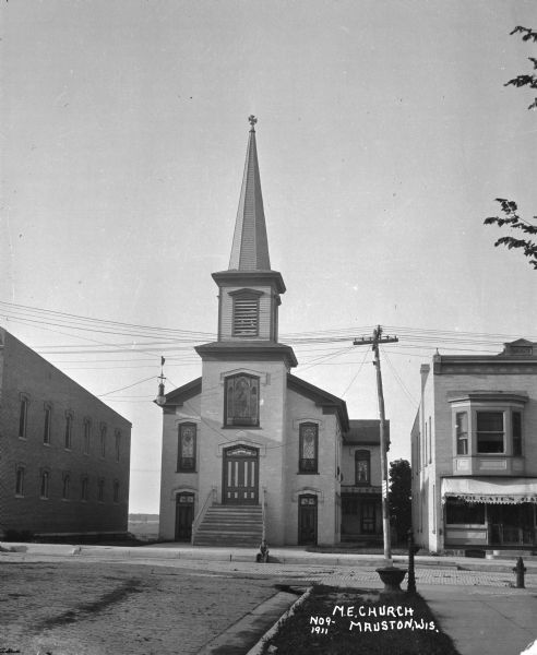Exterior of the Methodist Evangelical Church. A small boy sits on the curb along the cobblestone street outside the building. Next door to the church is Holgate's barbershop.
