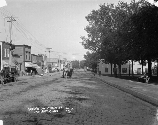 View down brick paved Main Street. A horse and carriage travels along the street. Caption reads: "Scene on Main St., Mauston, Wis."