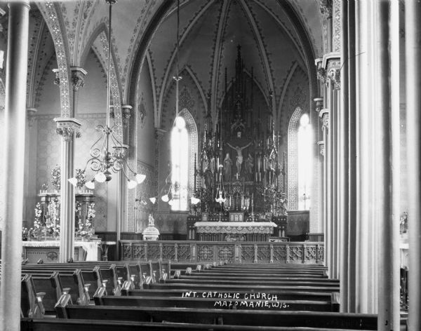 Interior of the Catholic Church looking over the pews towards the altar. Chandeliers hang from the arched ceiling, and columns are along the sides of the pews.
