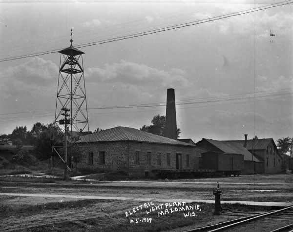 The site of the Electric Light Plant located near railroad tracks. There is a bell tower with a weather vane on the left.