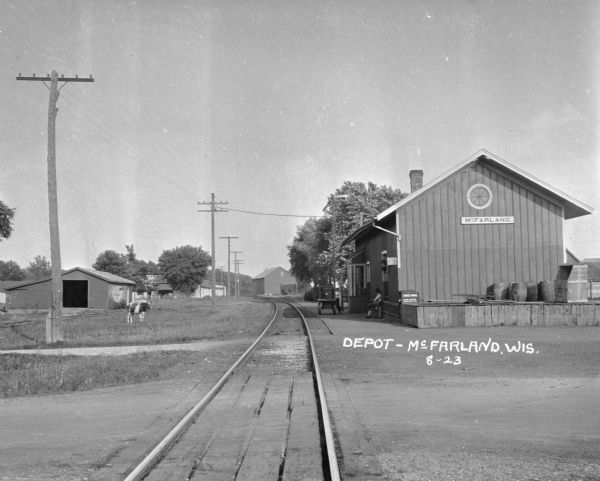 View down railroad tracks of the McFarland train depot. Two men are on the platform, and a cow grazes in the grass on the left.