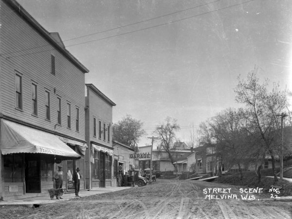 View down the curved unpaved street. Two men and a dog stand under the awning of a shop on the left. Another group of men are posed near a car parked outside of an automotive repair garage.
