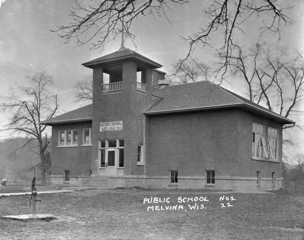 Exterior of the first district public school, erected in 1916. There is a hand-pump for water in the yard in front of the building.