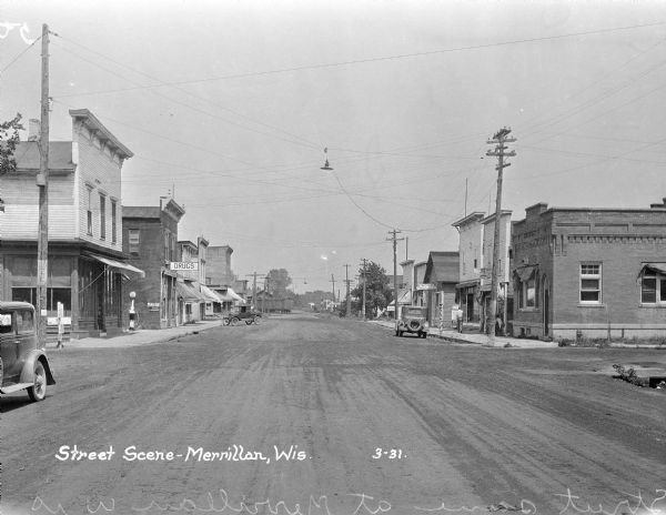 View down a commercial street. The shops on the street include: a drugstore, the City Hotel, the post office, the Merrillan Home Bakery, and the State Bank. At the end of the street is a railroad car.