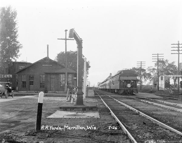 View along railroad tracks towards the Merrillan depot and train. The Hotel Campbell is located on the left. A group of children and adults are sitting outdoors at the end of the last railroad car of a train. Pedestrians stand on the platform.