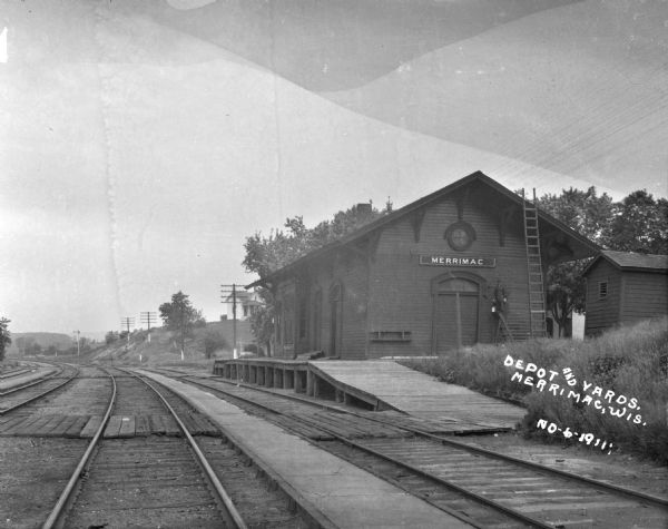 View across railroad tracks of the Merrimac train depot. Against the building a man poses on top of a ladder. He is repainting the exterior. There is a house on a low rise in the background.