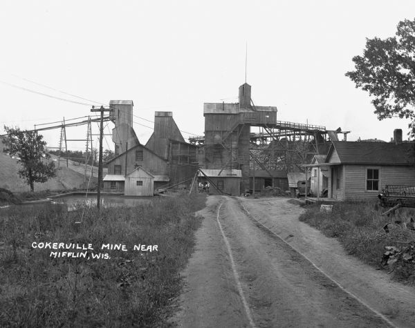 View down road leading up to the Cokerville Mine. Two horses pulling a wagon are near the road, and automobiles are parked on the right near a building with a porch.
