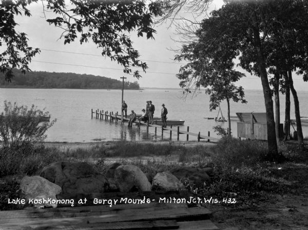 View from shore of a group of men posing on a dock at Burgy Mounds on Lake Koshkonong. Boats are tied to the dock. There are a number of people in boats far out on the lake near the far shoreline.