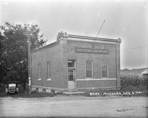 Exterior of the Bank of Mindoro. A car is parked on the lawn beside the bank. On the back of the car is an advertisement for the Interstate Fair in La Crosse. On the right is a crop of corn in a field.
