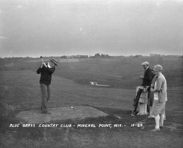 Two men and two women play a game of golf at the Blue Grass Country Club. One man tees off as the other three players watch.