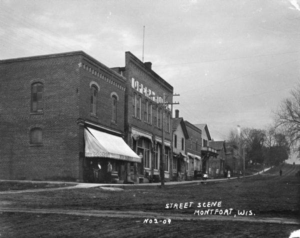 View from unpaved street of a business district. W.G. Baxter's dry goods store is on the corner, and next to it is the Opera House. A number of people are standing or walking along the sidewalk. In the background the street rises up to a hill.