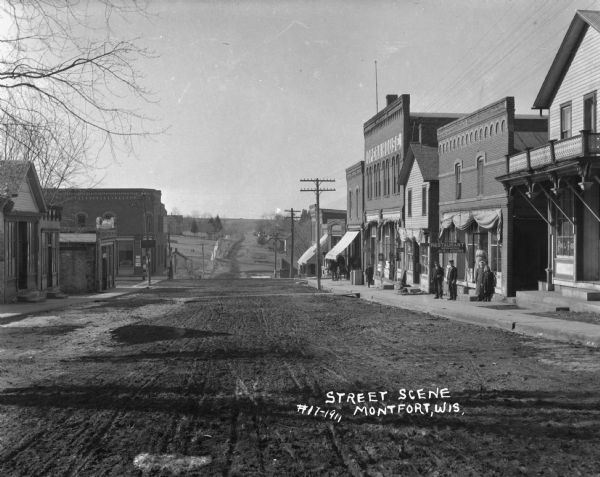 View down an unpaved street in the business district. The buildings along the street include: a pool room, restaurant, opera house, W.G. Baxter's dry goods store, and the State Bank. A number of people are posing on the sidewalk. The street drops down a steep hill starting at the State Bank, and fields and houses can be seen in the distance.