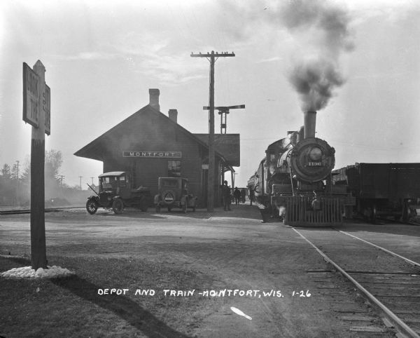 View across road along railroad tracks of a train departing the Montfort depot. Two cars are parked at the depot, and a number of people are on the platform. A sign on the left says: "Look out the Cars."