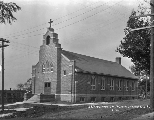Exterior of Saint Thomas Church. The brick building features a steeple, arched windows, and double doors. A plaque on the front says: "St. Thomas 1925."