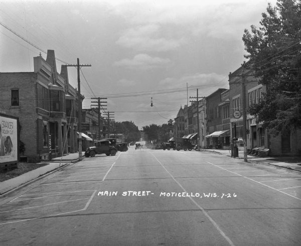 View down Main Street of cars and shops lining both sides of the street. The buildings include: the Grand Central Hotel, an ice cream shop, and an automotive repair and filling station. A barber  pole is in front of the first building on the left.