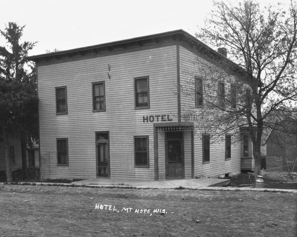 A hotel on an unpaved road. A man stands looking out of the hotel from behind a screen door at the corner of the building.