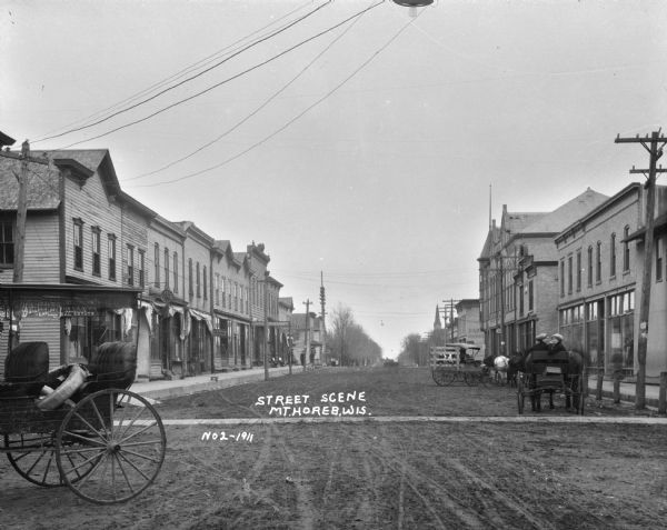View down street lined with businesses. There is a surrey or carriage with fringe along the top in the foreground on the left, and two women sit in a horse-drawn vehicle on the right. A number of other carriages and automobiles are parked along the sidewalk. The shops include: a dentist's office, City Meat Market, and insurance and real estate office and an automotive garage.