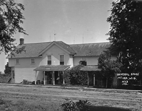 View across road of the Mount Ida post office and general store. A man is sitting on the porch.