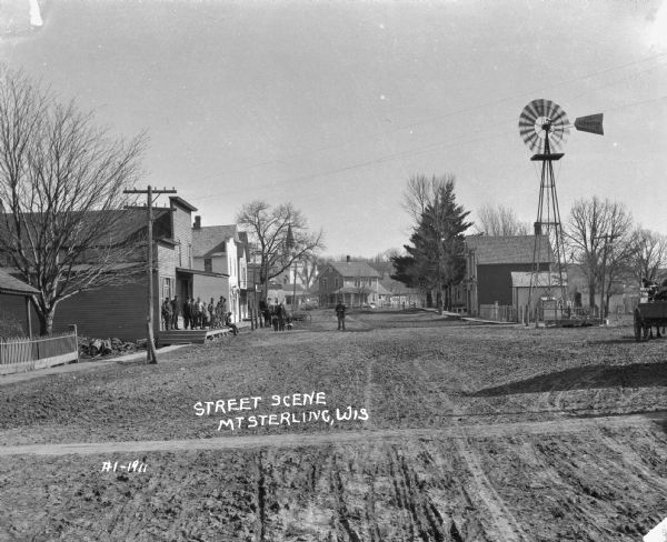 View down unpaved street. A group of men and boys congregate on the porch of a storefront. Two men and two children stand in the street. There is a windmill with a hand-pump underneath it on the right.