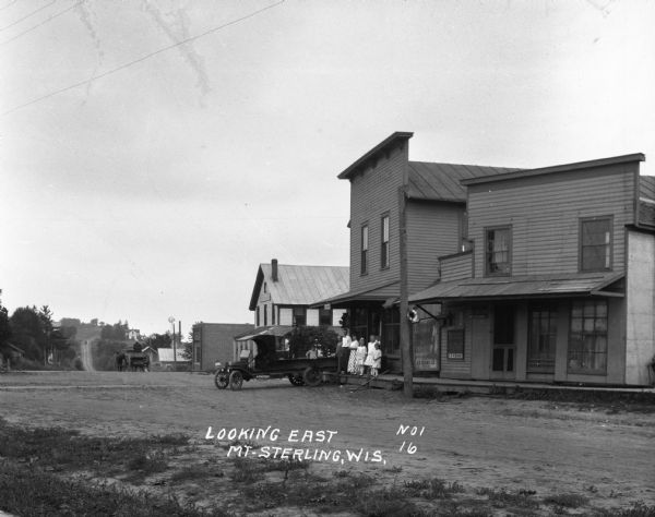 View down unpaved street. A group of men, women, and children pose on the porch of a post office. Near them a man and boy pose on a pickup truck which is parked with the bed over the porch. The road dips down and up a hill where there is a house and windmill. A man is driving a horse-drawn wagon near a brick building.