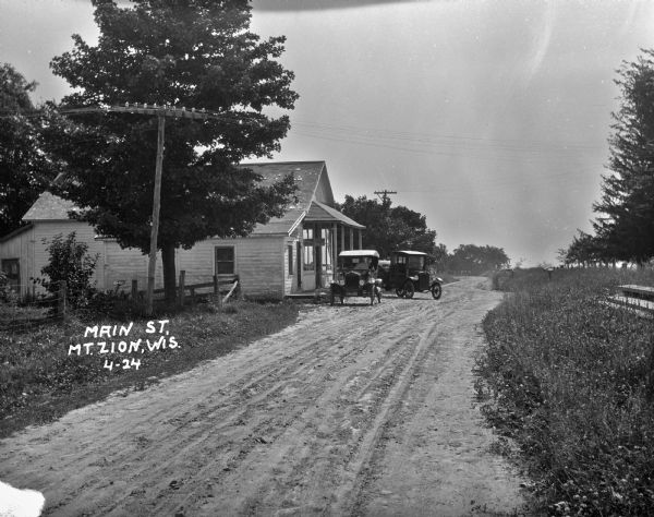 View down unpaved road of two automobiles parked outside of a store on Main Street.