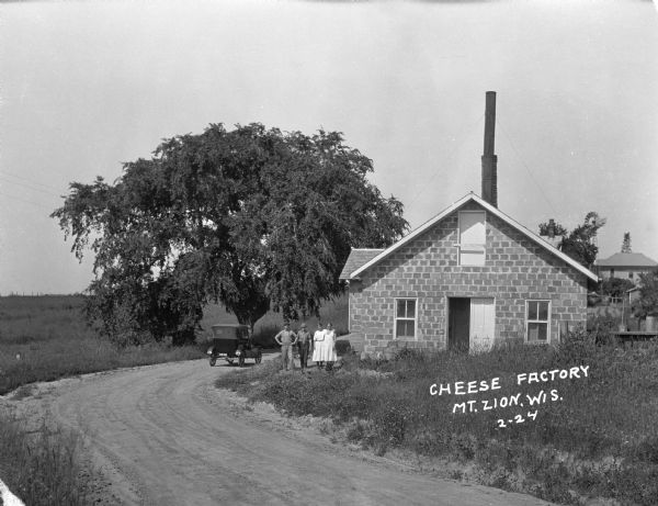View from unpaved road of two men and two women posing outside of a cheese factory. An automobile is parked in the road near a large tree. In the background is a house and a windmill.
