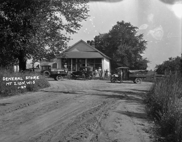 View down road of the exterior of a general store. Men, women, and children pose near the three parked vehicles outside the storefront. One of the vehicles is a truck with signs for "Standard Oil Company / Red Crown Gasoline."