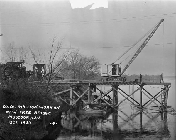 View along shoreline of construction of the New Free bridge. Men work on and below the wooden bridge, which is partially constructed. A crane is on the bridge to assist in the construction.
