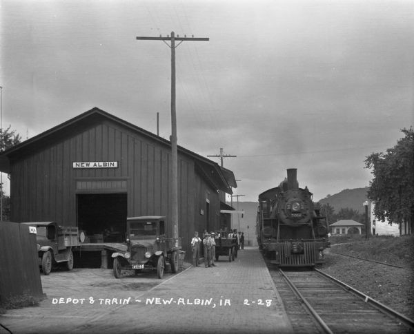 View down cobblestone platform of a group of young boys and men standing alongside the front of the depot. The locomotive on the tracks is Class A1 3012, an engine of the Atlantic (4-4-2) type, of the Milwaukee Road. Trucks are parked at the loading dock at the side of the depot on the left. There are bluffs in the background.