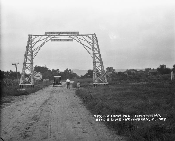 View down road of a man and young girl posing near their car at the Iowa welcome sign on an archway over the road. In the background are advertisements for Red Crown Gasoline and Star Brand Shoes. On the far right along the roadside is an iron post noting the state line separating Iowa and Minnesota.
