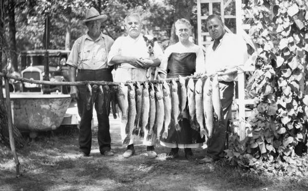 Three men and a woman pose with a pole of a stringer of fish. One of the men holds his pet dog. Behind them on the left is a bathtub, hand-pump for water, and an automobile.