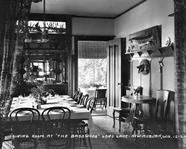 Dining room at the Basswood in Long Lake. Fish are mounted on the wall on the right, and along the back wall is a china cabinet with stained glass above. Through a doorway in the background, there appear to be more tables on a porch.
