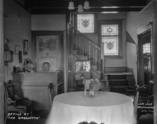A man works at the front desk at the lobby of the Basswood resort in Long Lake. The room features a table and chairs, stained glass windows, a calendar, and a telephone.