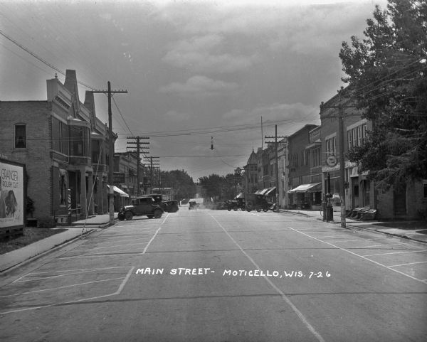 View down Main Street. Shops and parked cars line the street. On the  near left, there is a billboard for Granger rough cut tobacco, and there is a barbershop pole nearby on the sidewalk. Shop signs include: the Grand Central Hotel, an ice cream shop, and Monticello Automotive.