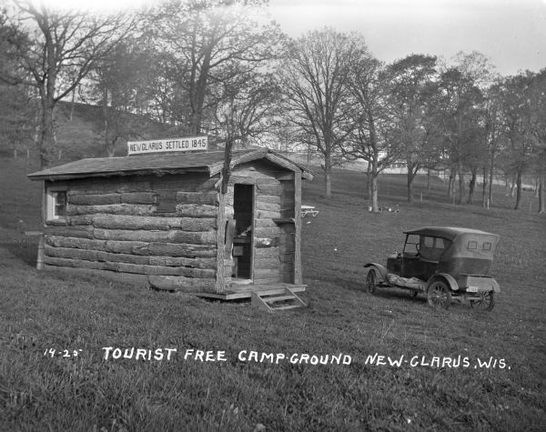 A log cabin and park car at the Tourist Free Camp Ground. The cabin has a sign that reads, "New Glarus Settled 1845."