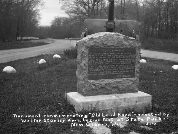 A stone monument dedicated to the Old Lead Road. The marker reads: "The Old Lead Road. In 1828, ox-teams, guided along an ancient Winnebago Indian Trail, began hauling over this road. June 29, 1832, after the battle of the Pecatonica, Gen. Henry Dodge and his Rangers passed here to join the main pursuit of Black Hawk at Koshkonong. June 27, 1845, Niklaus Durst and Fridolin Streiff were led by this road to the site of New Glarus which they selected as the location of their Swiss colony. August 15, 1845, the first band of Swiss colonists followed this road to their new home. Erected by Walter Stuessy, Post No. 141, American Legion".