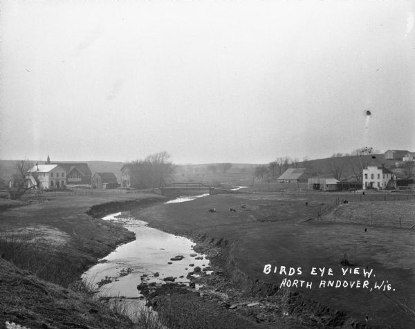 View from low hill of a stream flowing through fields. There are cows in the pasture on the right. In the distance is a bridge, road, and buildings.